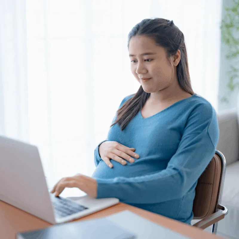 Pregnant woman working on a laptop at her desk, one hand on her belly