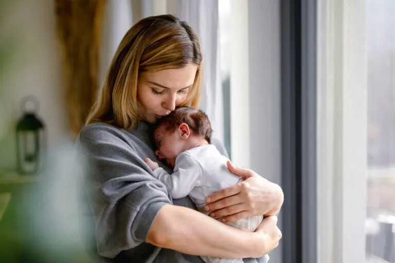 Mother in a grey sweater kissing the head of the newborn she holds against her shoulder