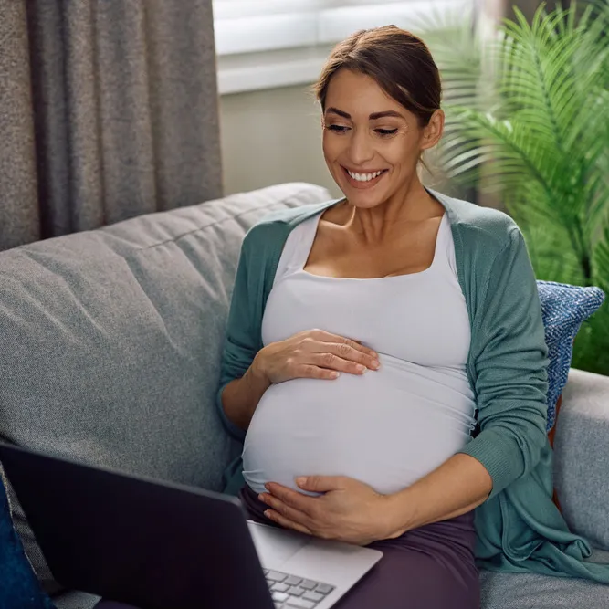 Smiling pregnant woman on a sofa with her hands on her belly and a laptop beside her