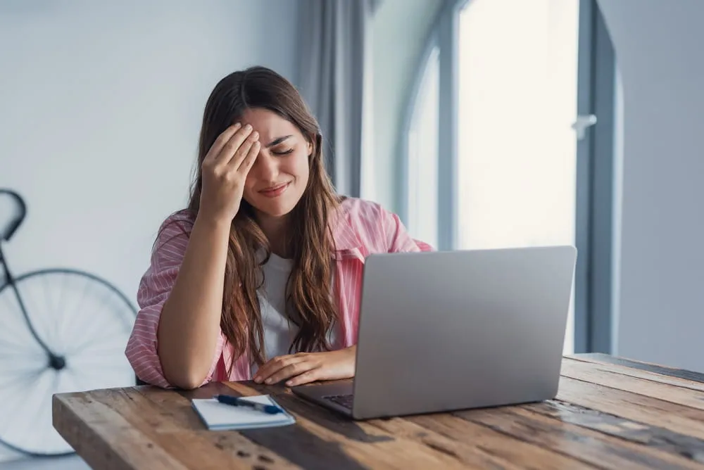 Young woman at a laptop, holding her forehead and smiling as if overwhelmed