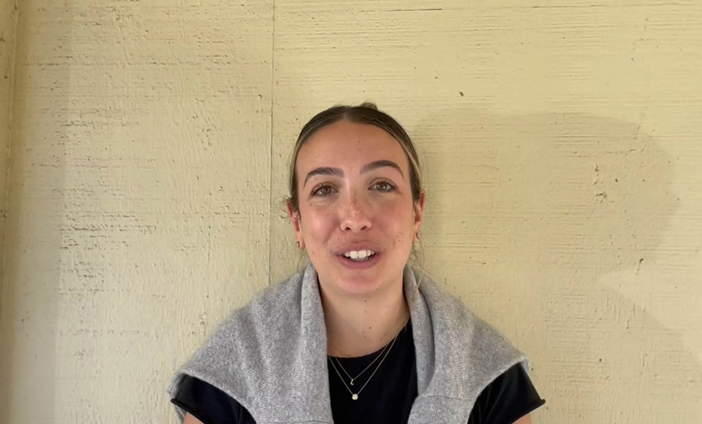 Young woman with a grey sweater over her shoulders, speaking to the camera